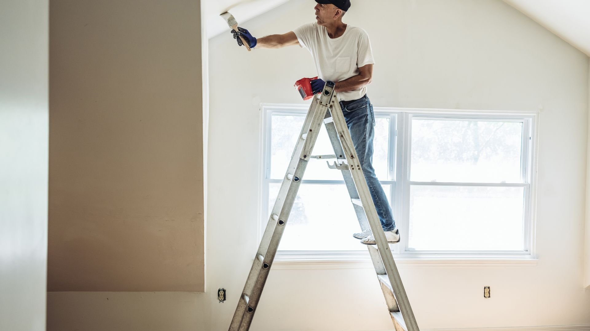 Person painting wall while standing on ladder near windows